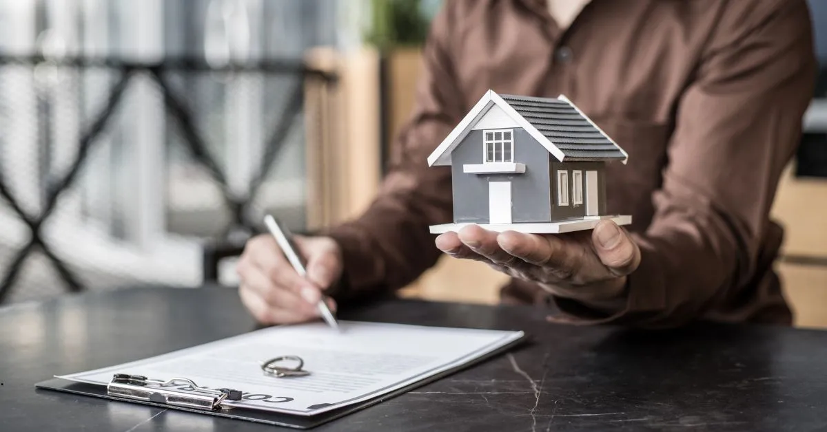 Pessoa segurando uma miniatura de casa enquanto assina um contrato de compra e venda de imóvel.