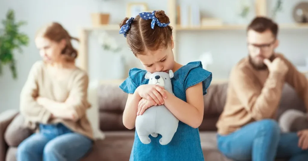 Criança triste abraçando um urso de pelúcia enquanto os pais discutem sobre guarda unilateral ao fundo.
