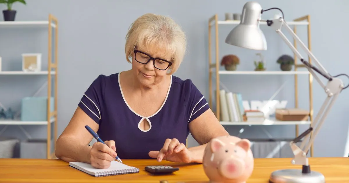 Mulher idosa fazendo cálculos com caderninho e cofre de porquinho ao lado, representando planejamento previdenciário