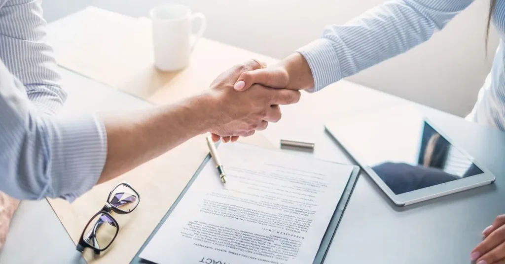 Duas pessoas apertando as mãos em cima de um contrato de locação assinado, simbolizando acordo de aluguel.