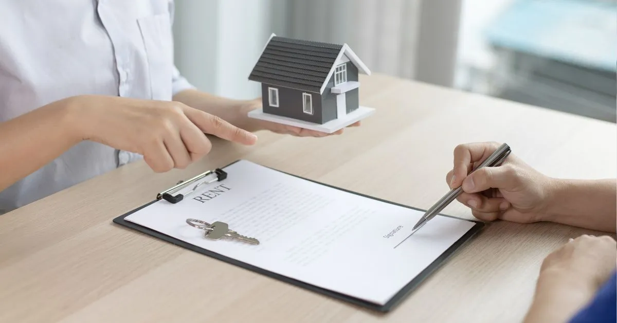 Pessoa assinando contrato de aluguel com chave e maquete de casa na mesa, representando a responsabilidade do fiador em caso de atraso.